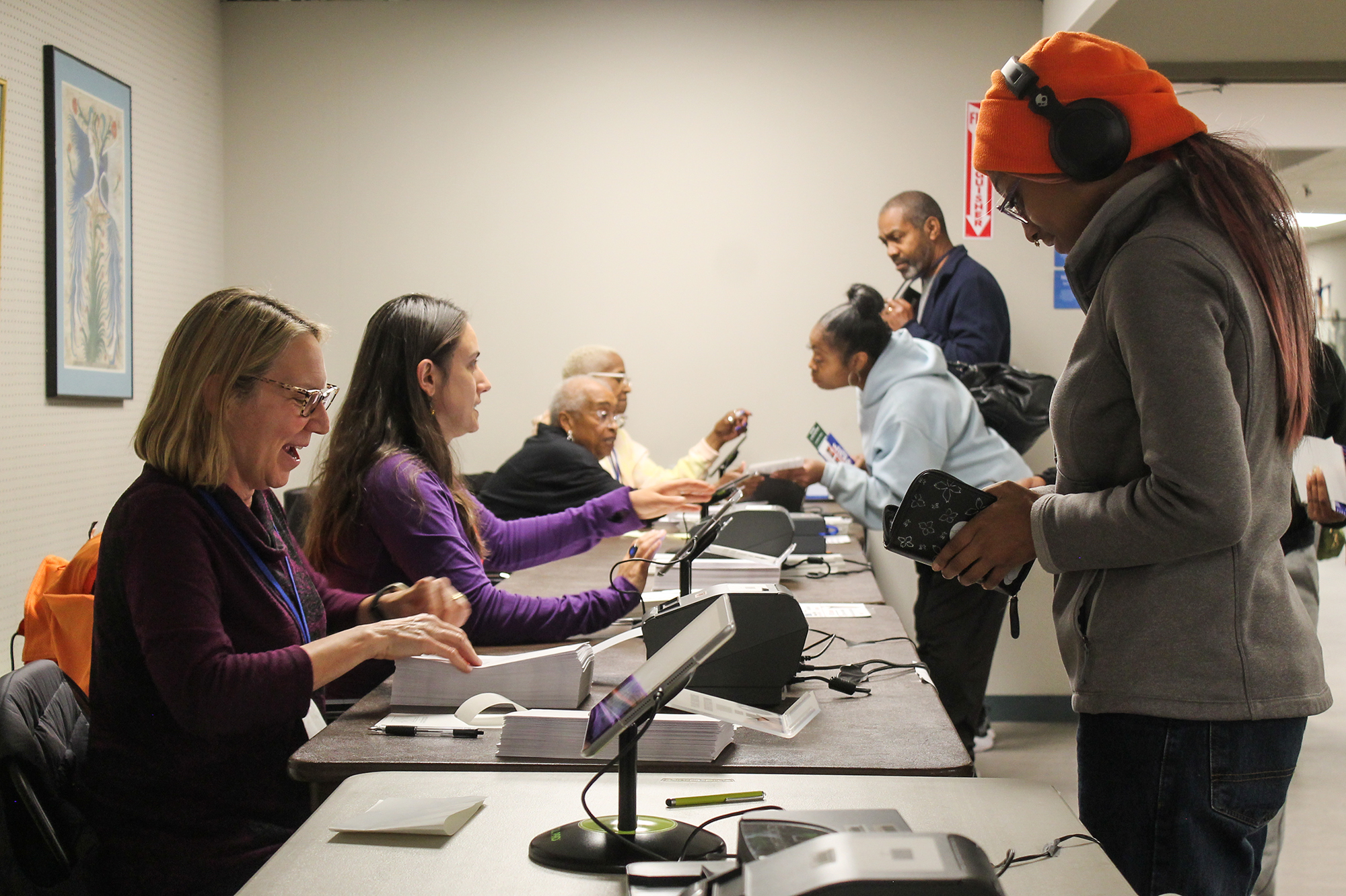 A middleaged White woman smiles as she interacts with a small tablet. In front of her is a young Black woman who wears an orange hat over braided hair. She is a first-time voter.